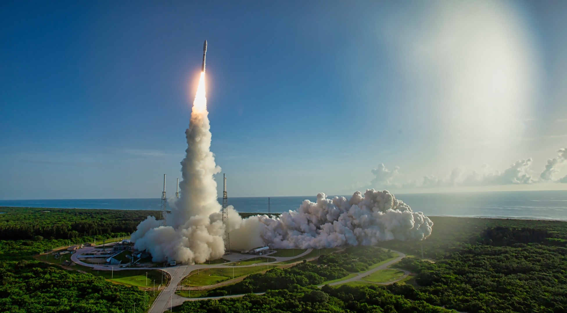 A rocket lifts off from a coastal launch pad with bright exhaust plume and a smoke trail rising into a blue sky